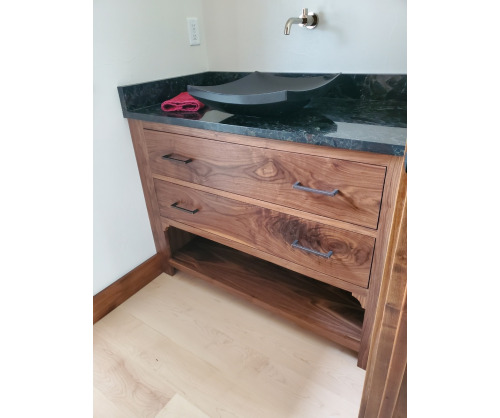 Wide vanity with 2 large drawers and a open shelf on the bottom with a dark stone countertop and artistic raised sink with a wall mounted faucet